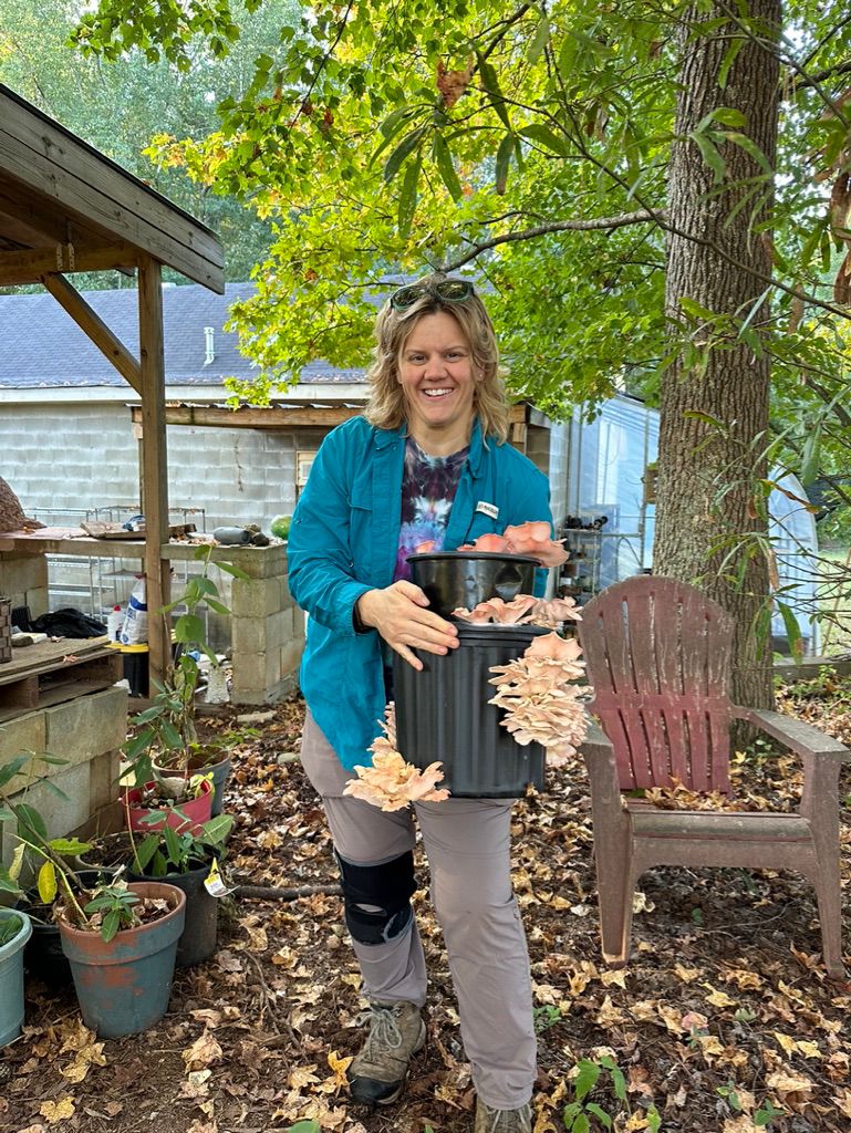 A person with medium length hair holding two black buckets containing fruiting mushrooms